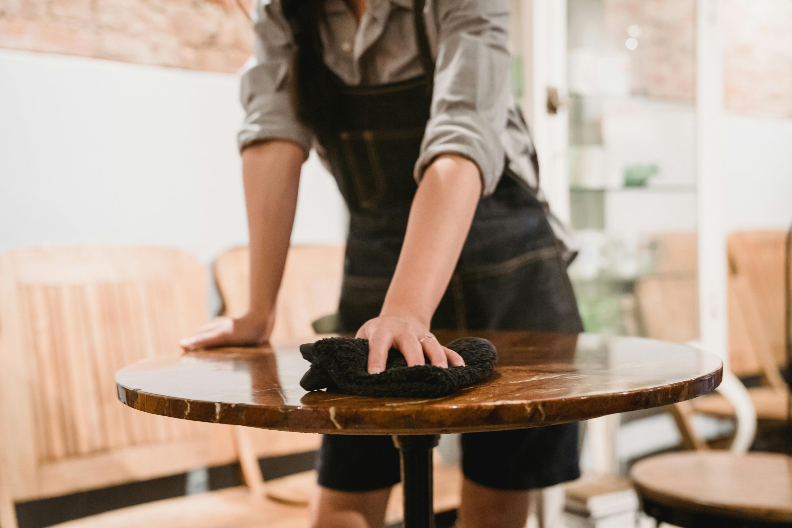 Person in apron cleaning a wooden table in a cozy café environment, focusing on hygiene.