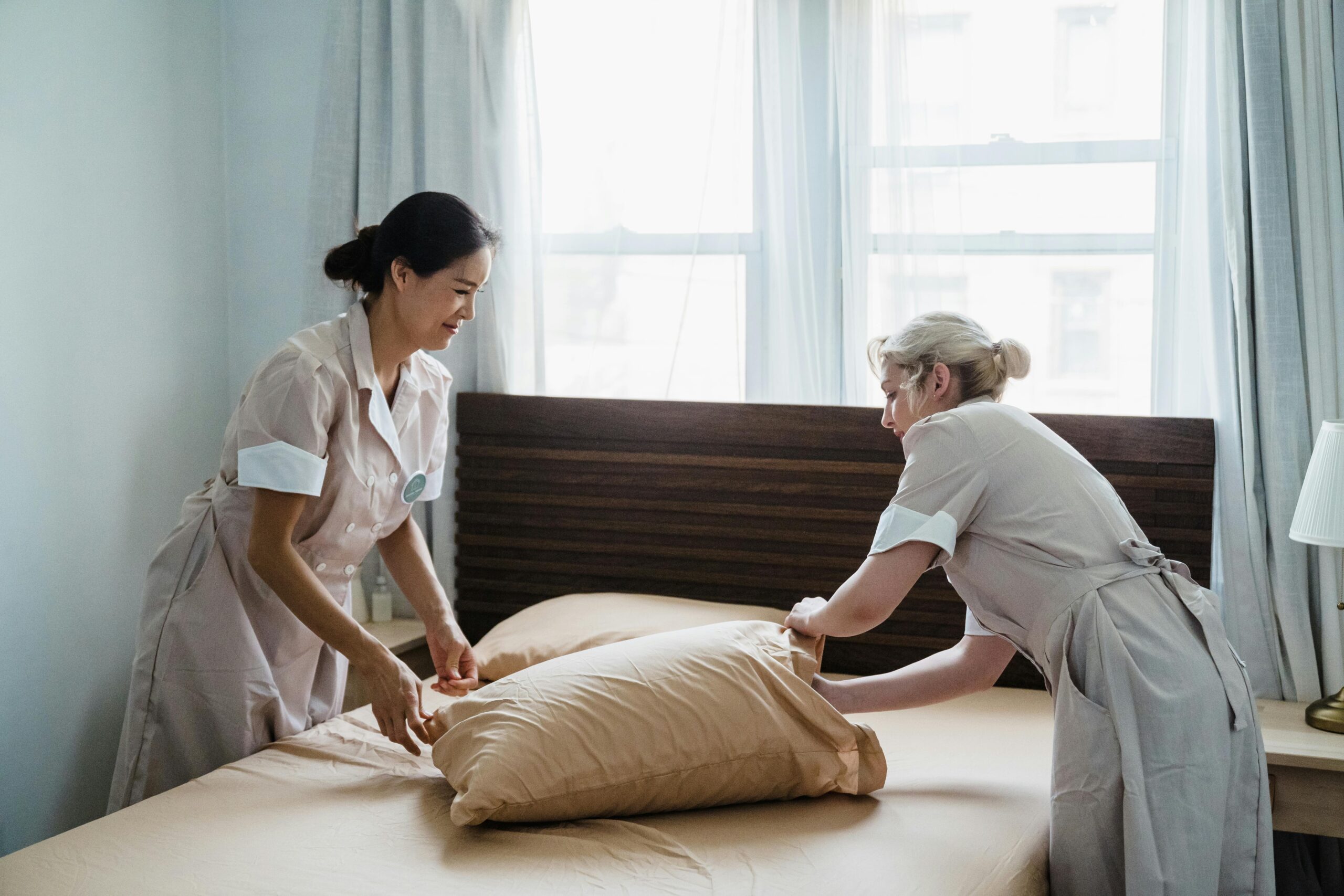 Two housekeepers in uniform collaborate while making a bed in a bright, peaceful bedroom.